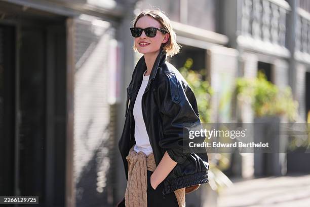 Guest wears sunglasses, a black leather jacket, a white cotton T-shirt, black wide leg trousers, a brown leather bag, a brown braided wool pullover,...