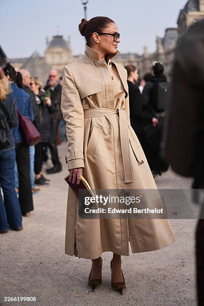 Guest wears brown hair in a bun, black sunglasses, a beige cotton belted trench coat, a burgundy leather clutch bag from Elie Saab, brown satin...