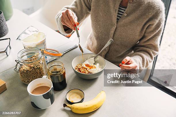 view of a mature woman eating a healthy homemade breakfast of granola, yoghurt and honey - ontbijt stockfoto's en -beelden