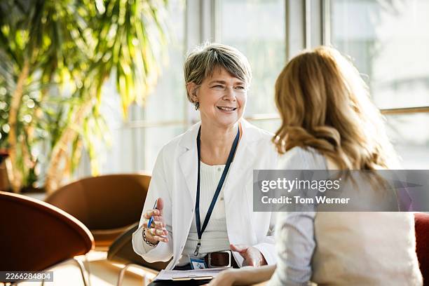 female mature healthcare professional explaining medical information to female mature patient in bright clinic lounge - zorgverzekering stockfoto's en -beelden