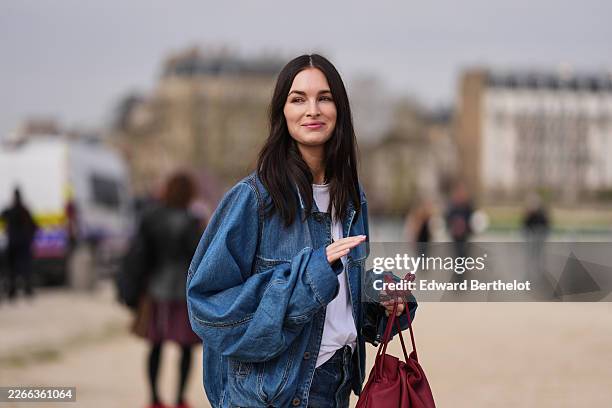 Guest wears dark brown hair, a blue denim jacket, a white cotton T-shirt, blue denim bootcut jeans, a red leather handbag, outside Loewe, during...