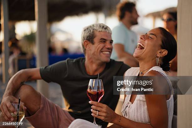 young adult couple enjoying drinks laughing at beach bar - outdoor dining stock pictures, royalty-free photos & images