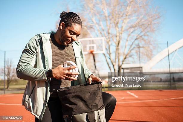 smiling young man of african ethnicity kneeling with sports bag on court - gym bag stock pictures, royalty-free photos & images