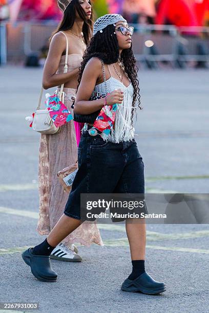 Festival-goer wears silver grey crochet knit fringe crop top with chain straps, black washed wide-leg denim bermuda shorts, large silver concho belt,...