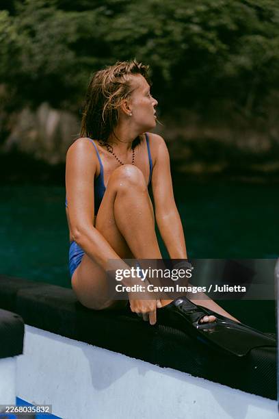 woman in swimsuit putting on diving fin while sitting on boat - schwimmflosse stock-fotos und bilder