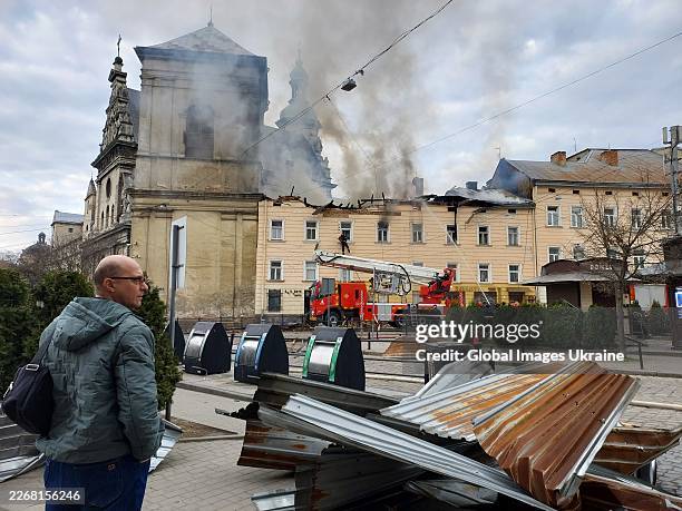 Man stands on the walk side while firefighters extinguish a fire on building in center of city after Russian drone attack on March 24, 2026 in Lviv,...