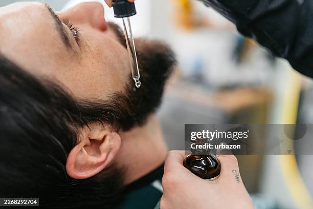 young female barber putting aftershave oil on a young man's beard at a barber shop - facial oil stock pictures, royalty-free photos & images