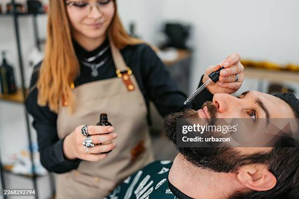 young female barber putting aftershave oil on a young man's beard at a barber shop - facial oil stock pictures, royalty-free photos & images