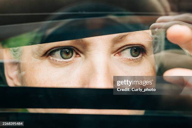 a dramatic close-up of a woman's eyes peering through dark horizontal blinds. - fobia imagens e fotografias de stock
