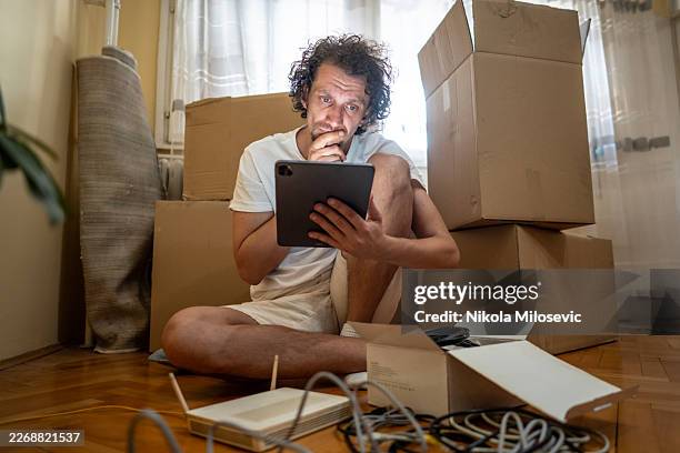 thoughtful man using tablet while surrounded by moving boxes during apartment move and unpacking - sitting on ground stock pictures, royalty-free photos & images