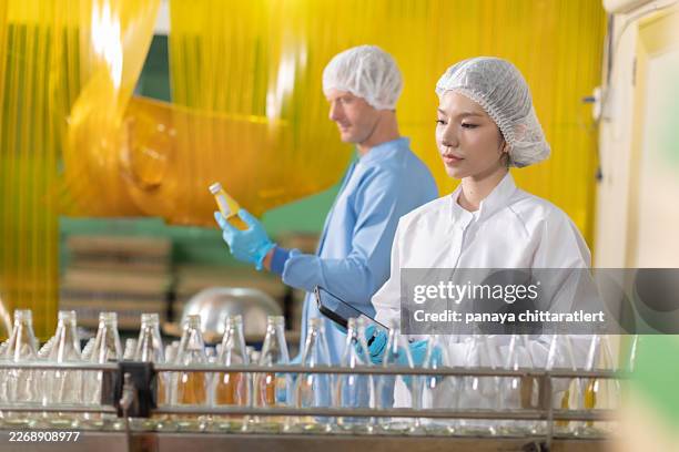 young caucasian man worker and asian female manager discussion.scientist worker checking the quality of machine system at the industrial factory. .asian female worker inspect the quality of bottled fruit juice products. - bottling plant stock pictures, royalty-free photos & images