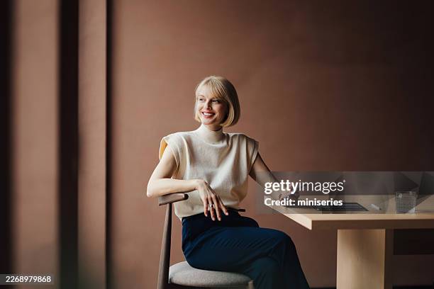 elegante empresaria sonriendo en una oficina moderna sosteniendo un smartphone junto a una tableta y un vaso de agua - presidente de organización fotografías e imágenes de stock