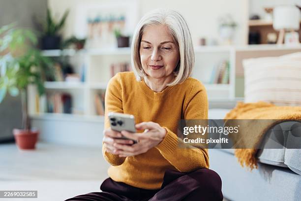 smiling mature woman with smart phone sitting on the floor at home - femininity stock pictures, royalty-free photos & images
