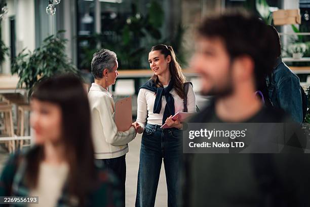 mature adult teacher advising young adult university student in modern campus building hallway during break time - generation gap stock pictures, royalty-free photos & images