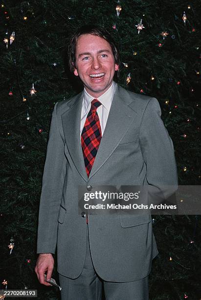 American actor John Ritter, wearing a grey blazer over a white shirt with a red-and-black plaid tie, poses beside a Christmas tree at an event,...