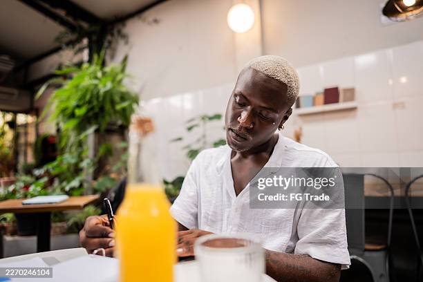 young businessman taking notes while working remotely in a coffee shop - shaved head stock pictures, royalty-free photos & images