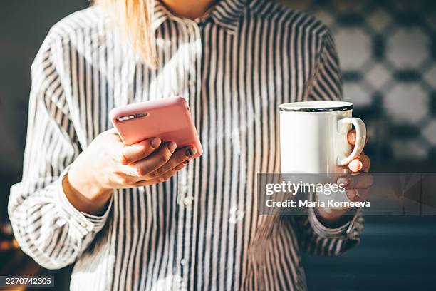 a person in a striped shirt holds a pink smartphone and a white mug in a bright kitchen. - mensagem com foto imagens e fotografias de stock