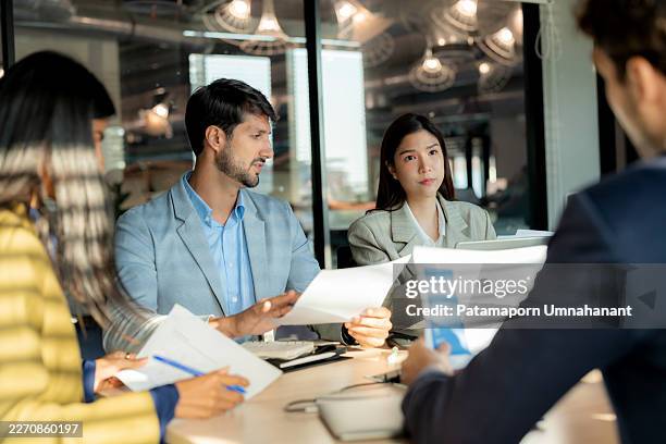 high-angle view of diverse executives analyzing and passing documents in a sunlit boardroom. a professional team collaborating on business strategy and paperwork during a bright afternoon in a modern corporate office. - businesswear stock pictures, royalty-free photos & images