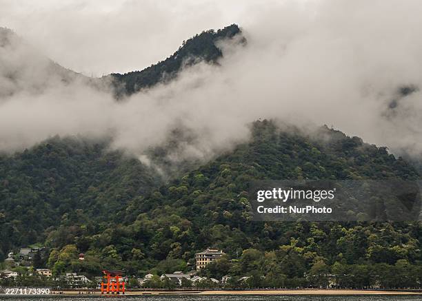 The iconic Great Torii Gate of Itsukushima Shrine stands before a backdrop of traditional inns and the forested slopes of Mount Misen on Miyajima...