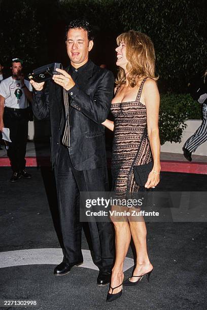 American actor Tom Hanks, wearing a black suit, inspects a Polaroid Vision Autofocus SLR camera , beside his wife, American actress and singer Rita...