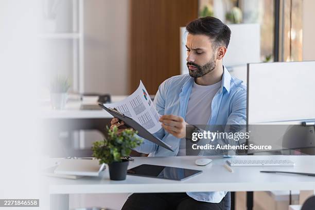 young professional man reviewing report at modern office desk while working on computer - proofreading stock pictures, royalty-free photos & images