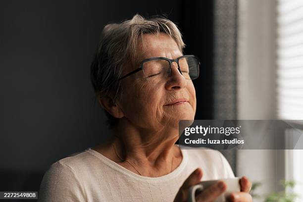 mujer mayor disfrutando de un tiempo tranquilo para ella en casa con una taza caliente y los ojos cerrados - escena de tranquilidad fotografías e imágenes de stock