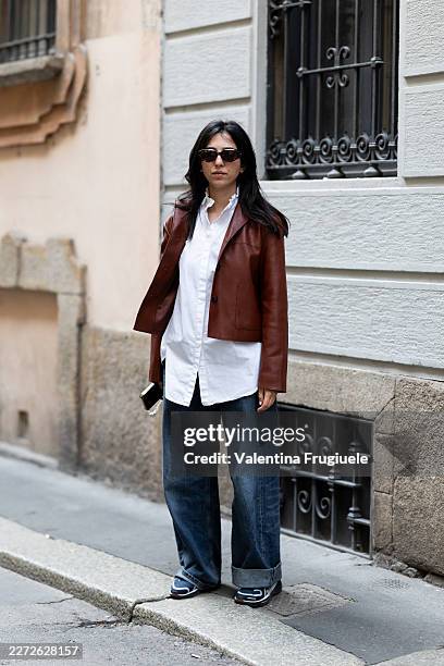 Passerby wears a brown cropped leather blazer over an oversized white poplin shirt, ultra-wide-leg dark wash denim jeans with a cuffed hem, dark navy...