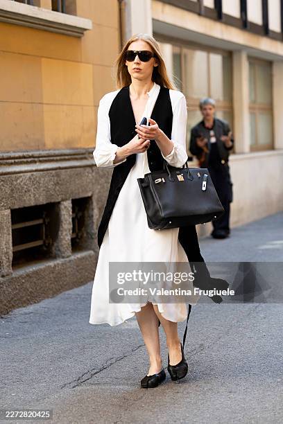 Passerby wears a long black sleeveless vest over a white fluid long-sleeve blouse, a white satin midi skirt, a black leather Birkin-style top-handle...