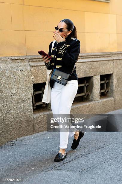 Passerby wears a navy military jacket with gold braid trim, white cropped jeans and black leather penny loafers, carrying a black Celine crossbody...