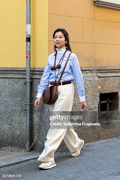 Passerby wears a light blue ruffle-collar shirt, cream wide-leg trousers and a brown leather belt, beige sneakers, carrying a brown leather saddle...