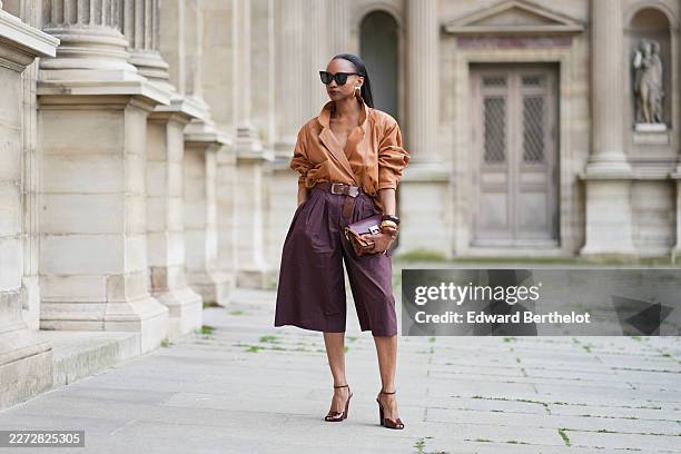 Emilie Joseph wears black Gentle Monster sunglasses, gold statement earrings, a brown vintage leather bomber jacket worn as a top, a brown leather...