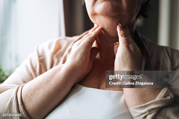 middle-aged overweight woman checking her neck and thyroid gland area with her hands by a home window, concept of self-examination and health awareness - schilddrüsenüberfunktion stock-fotos und bilder