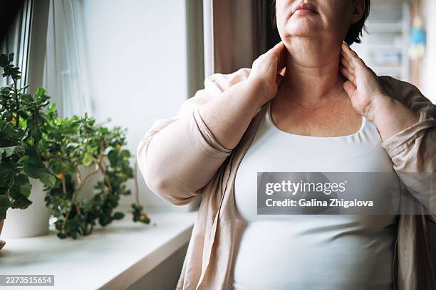 middle-aged overweight woman checking her neck and thyroid gland area with her hands by a home window, concept of self-examination and health awareness - schilddrüsenüberfunktion stock-fotos und bilder