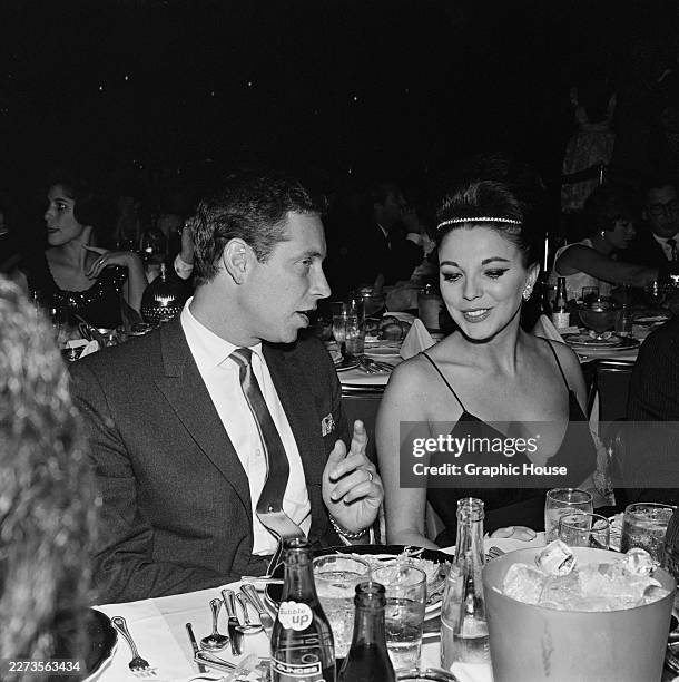 British actress Joan Collins, wearing a dark-coloured outfit with a plunging neckline and spaghetti straps, and Stanley Kamin, who wears a...