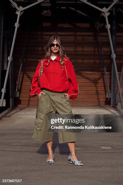Marina Lyritzi wears a red zip-neck pullover with white piping, olive green wide-leg cargo culottes, black sunglasses, carries a brown shoulder bag,...