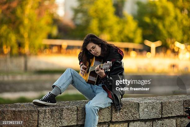 young woman playing acoustic guitar while sitting on a stone wall in a park at sunset - high tops stock pictures, royalty-free photos & images