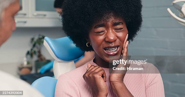 young woman with braces in dental chair holding cheek in pain during dentist visit - grimacing stock pictures, royalty-free photos & images