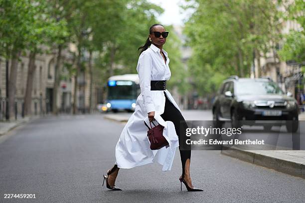 Emilie Joseph wears long straight black hair in a ponytail, black sunglasses, a gold-tone pendant necklace, a white longline Alaia shirt dress with a...