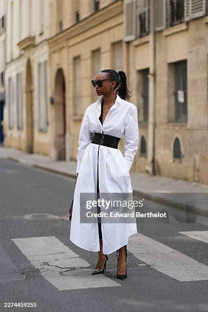 Emilie Joseph wears long straight black hair in a ponytail, black sunglasses, a gold-tone pendant necklace, a white longline Alaia shirt dress with a...