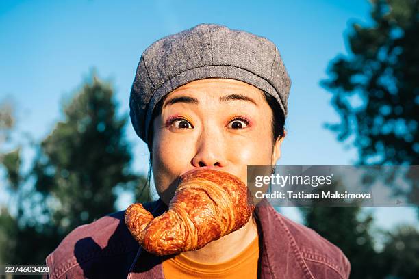 a colorful outdoor portrait shows a young person in a cap with a croissant, set against a bright blue sky and blurred trees, giving the image a quirky and playful editorial mood. - french food stock pictures, royalty-free photos & images
