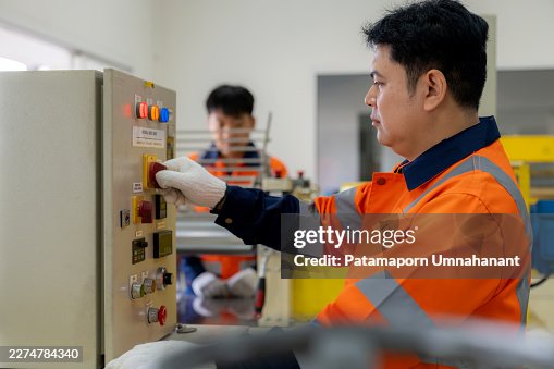 A mechanical engineer adjusting parameters on a control panel while a colleague operates a two-roll mill machine. Professional technicians managing polymer processing and rubber testing inside an industrial laboratory. A mechanical engineer adjusting parameters on a control panel while a colleague operates a two-roll mill machine. Professional technicians managing polymer processing and rubber testing inside an industrial laboratory.