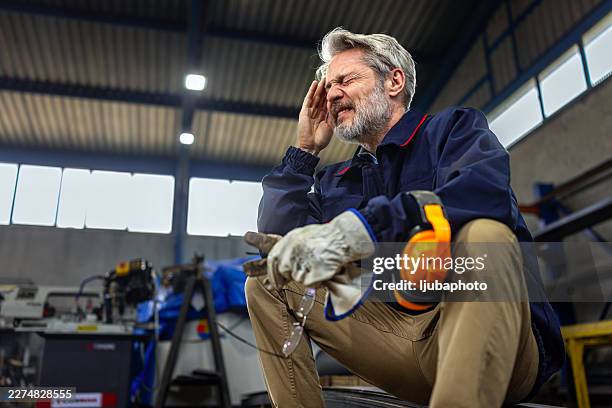 tired industrial worker in factory holding head with safety gear during metal manufacturing shift - safety equipment stock pictures, royalty-free photos & images
