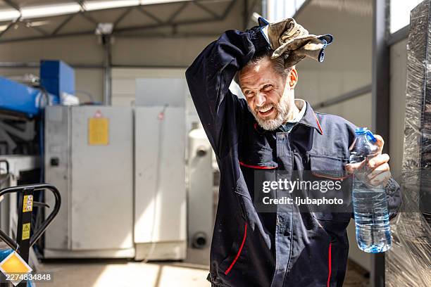 tired industrial worker taking break in factory holding water bottle after hot metal processing shift - overall stockfoto's en -beelden