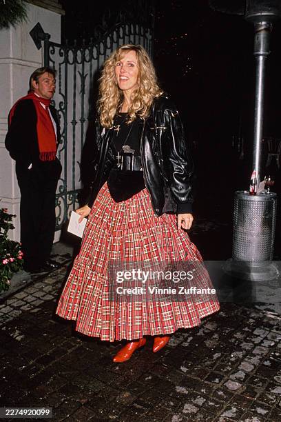 American actress Alana Stewart, wearing a black leather jacket over an evening gown with a black bodice and a red-and-white plaid full-length skirt,...