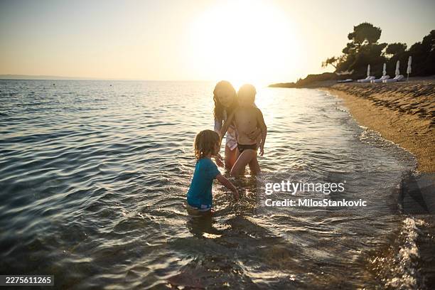 family children playing on beach at sunset — siblings splashing in shallow sea water - golden hour stock pictures, royalty-free photos & images