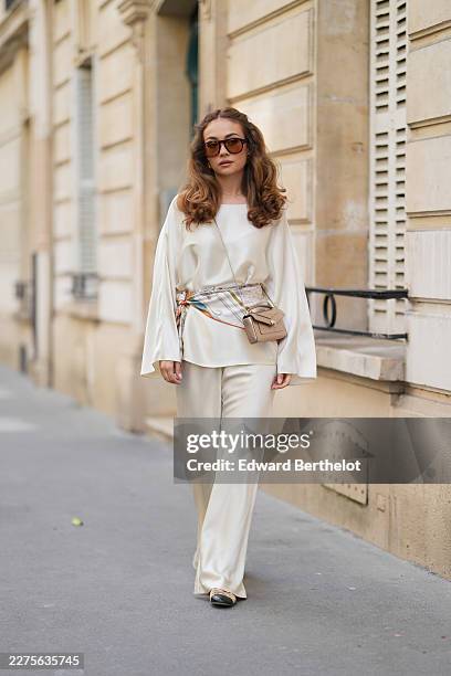 Merve Gorgoz wears long wavy light brown hair with a center part and voluminous curled ends, brown oversized Jimmy Fairly sunglasses with tinted...