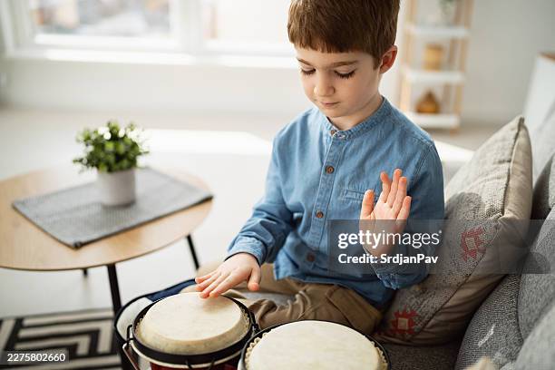 young boy practicing bongos on sofa in cozy living room - bongo drum stock pictures, royalty-free photos & images