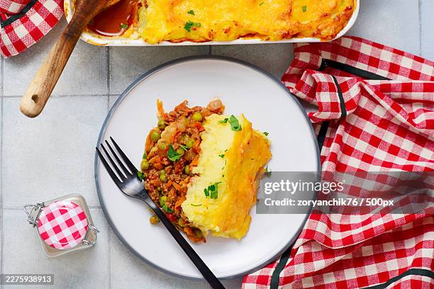 high angle view of a delicious shepherds pie serving on a plate with a full baking dish in the background, garnished with parsley and accompanied by a checkered napkin - cottage pie stock pictures, royalty-free photos & images