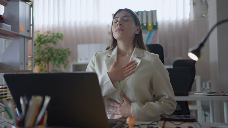 A woman in a white blazer sits at a desk with a laptop, eyes closed and hands on her chest and stomach, taking a deep, calming breath.
