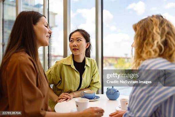 drei frauen, die sich am cafétisch mit tee und kaffee unterhalten - warmes licht stock-fotos und bilder